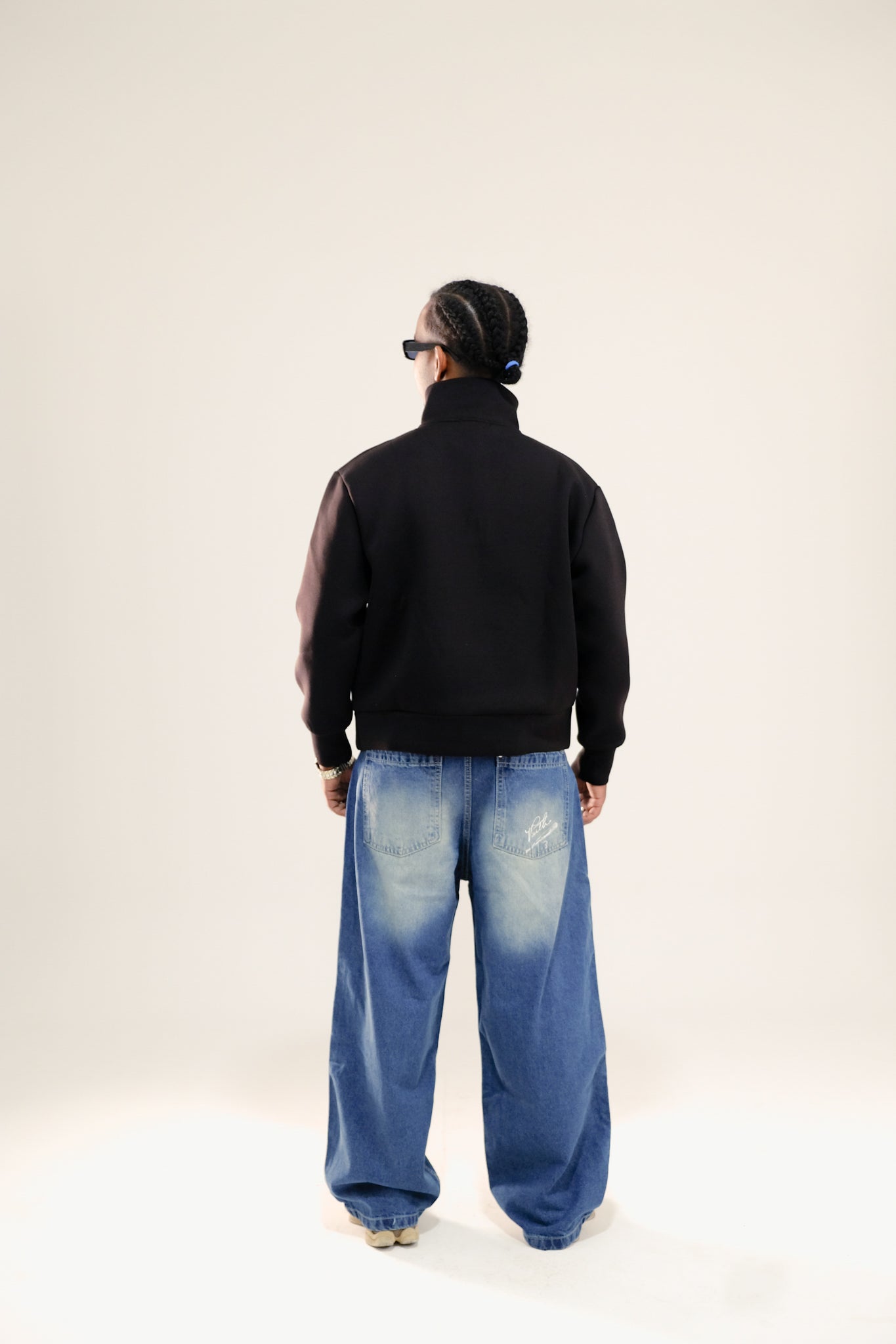 Man facing backwards wearing black quarter-zip sweatshirt and blue washed jeans standing against a beige background