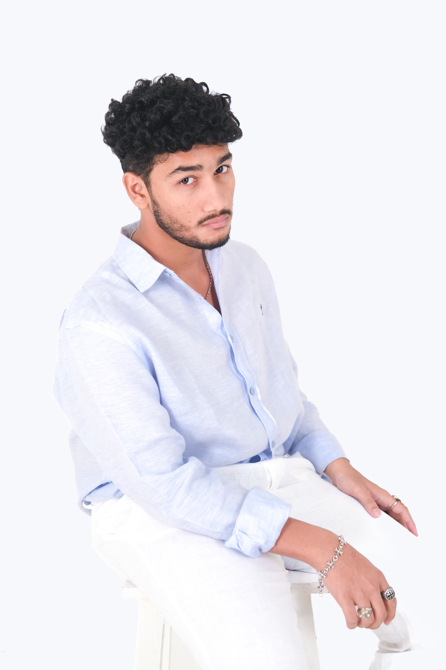 Male model seated on a stool wearing Version Originale’s classic blue linen shirt with navy palm tree embroidery, paired with Version Originale’s white linen pants