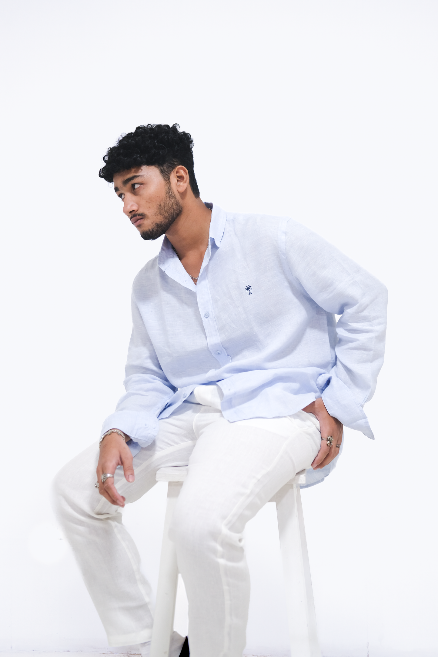 Male model seated on a white stool, styled in Version Originale’s classic blue linen shirt with navy palm tree embroidery, paired with Version Originale’s white linen pants
