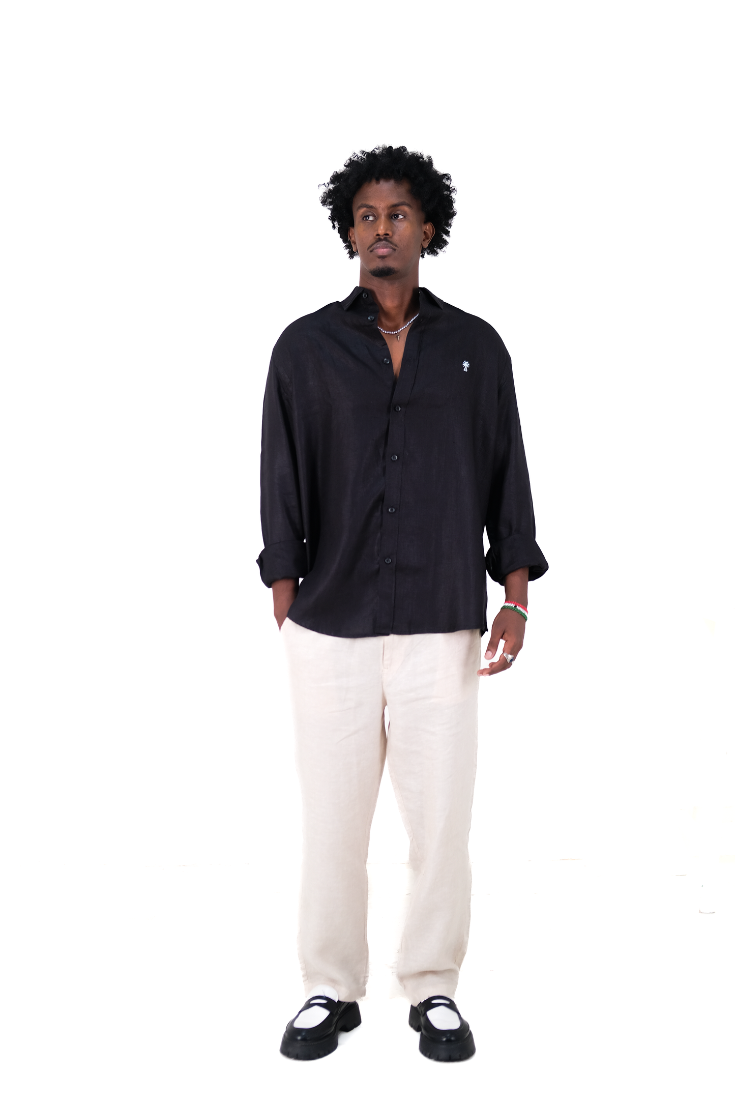 Male model wearing Version Originale’s black linen shirt with sky blue palm tree embroidery, paired with Version Originale beige linen pants and black loafers, standing against a white background