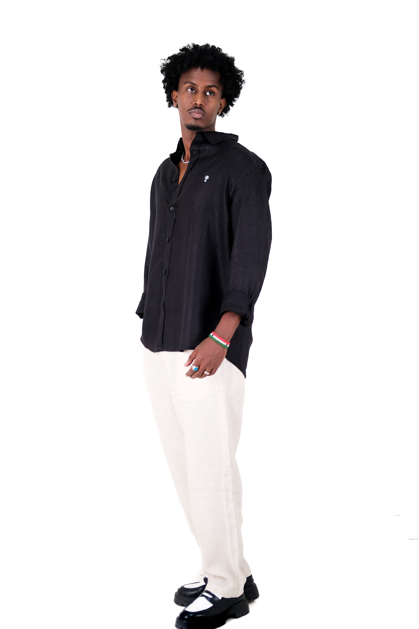 Male model wearing Version Originale’s black linen shirt with sky blue palm tree embroidery, paired with Version Originale beige linen pants and black loafers, standing against a white background