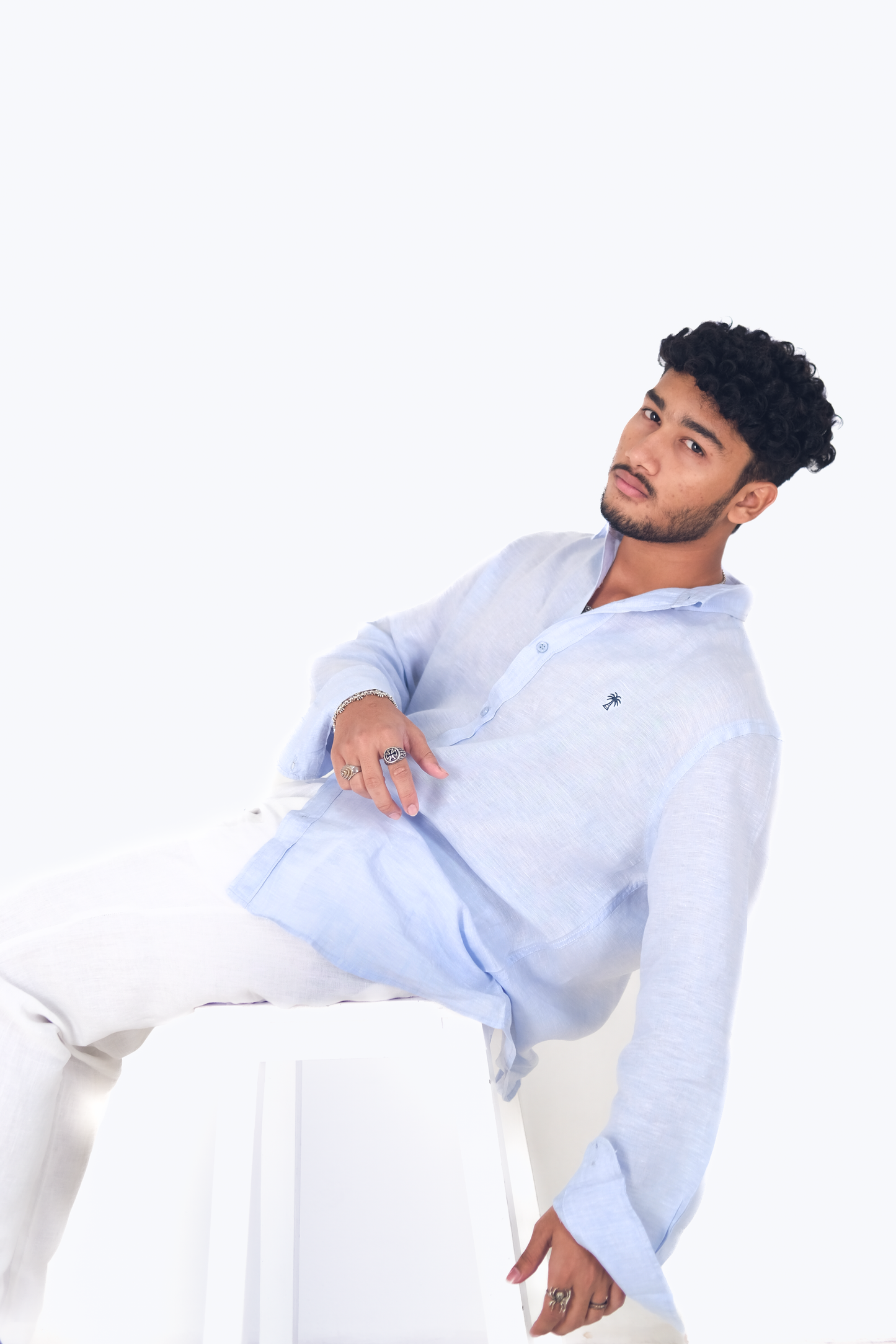 Male model lounging on a white stool, wearing Version Originale's classic blue linen shirt with navy blue palm tree embroidery and white linen pants, styled with silver rings and accessories against a light background