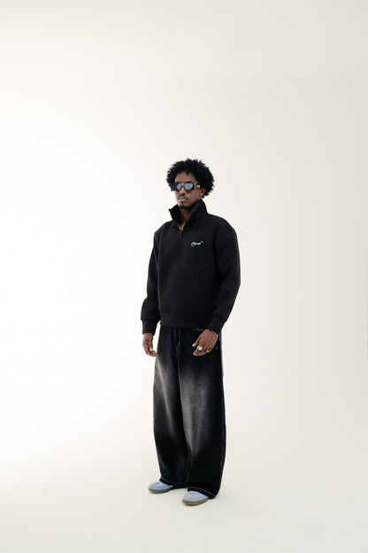 Man wearing black quarter-zip sweatshirt featuring a teal "Originale" embroidery and baggy black washed jeans standing against a beige background