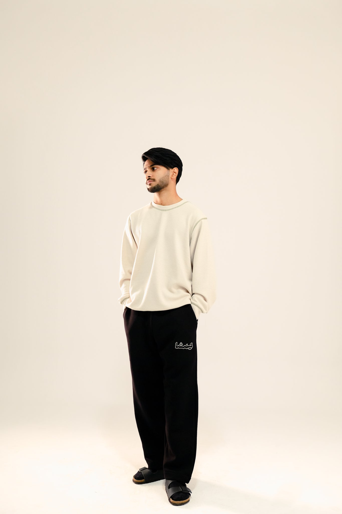 Man wearing a beige waffle knit sweatshirt and black sweatpants standing against a beige background