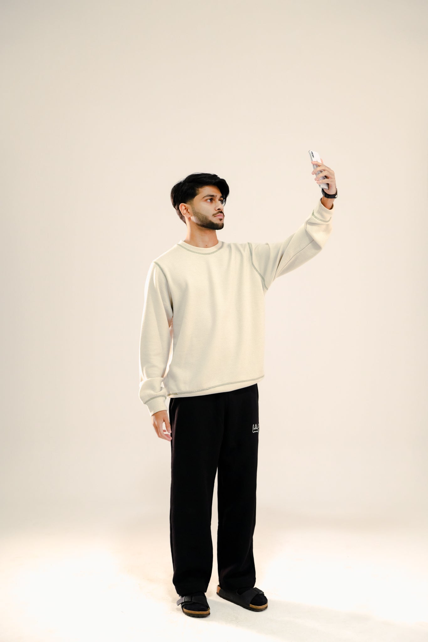 Man taking a selfie wearing a beige waffle knit sweatshirt and black sweatpants standing against a beige background