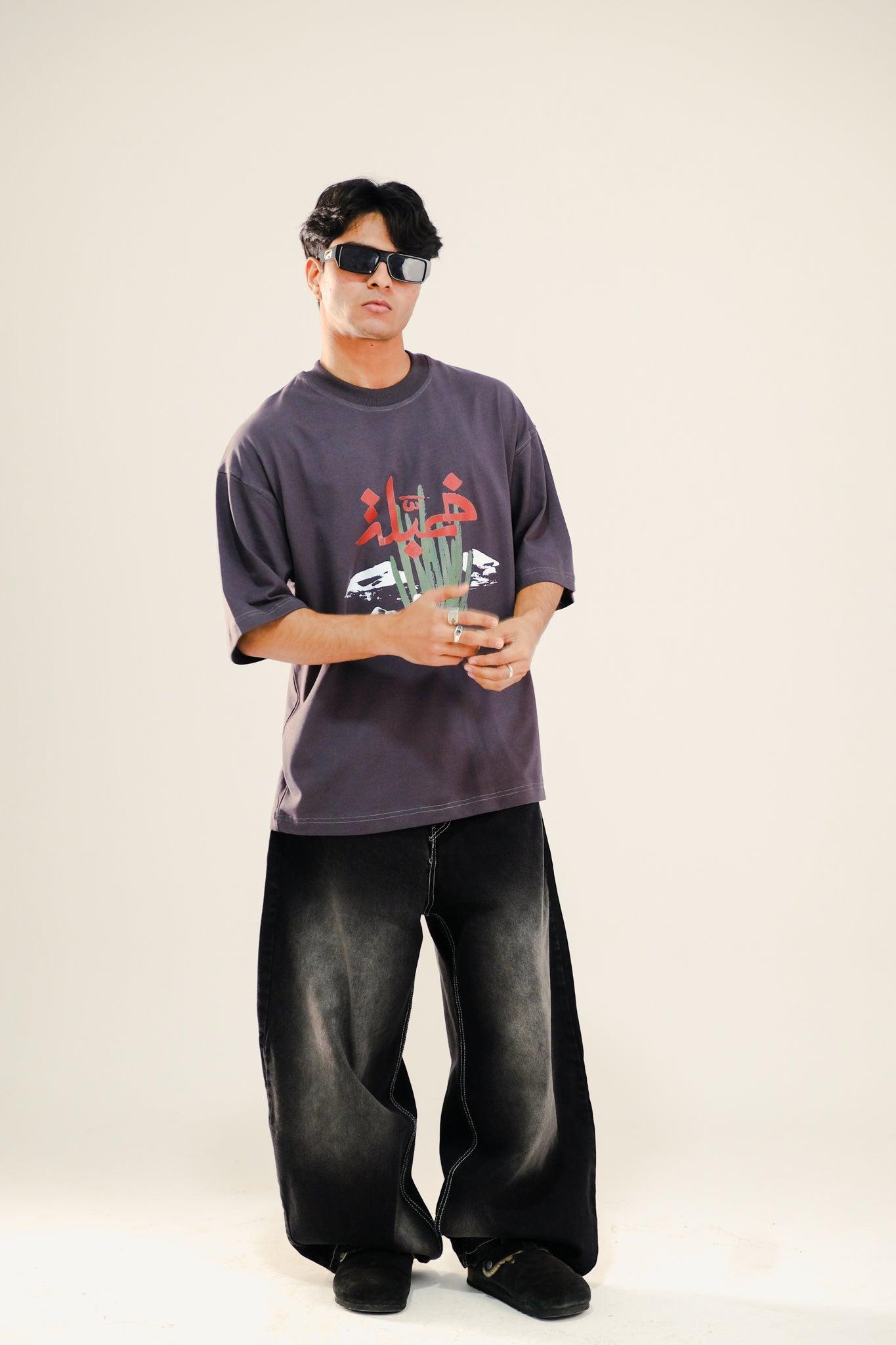 Man wearing grey drop shoulder t-shirt featuring an Arabian desert print and black washed jeans standing against a beige background