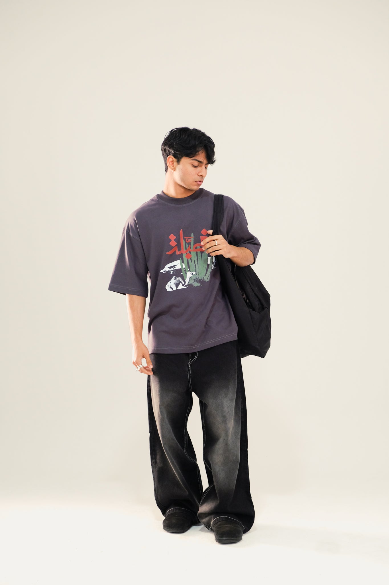 Man carrying a bag wearing grey drop shoulder t-shirt featuring an Arabian desert print and black washed jeans standing against a beige background