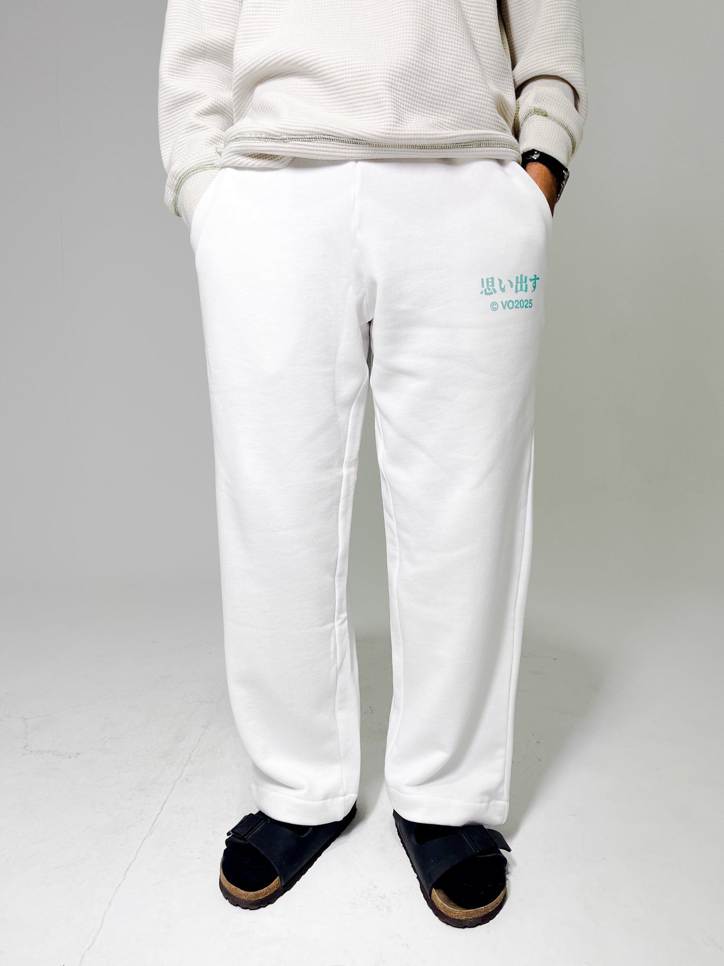 Model wearing beige sweatshirt and white sweatpants standing against a plain background with hands in pockets
