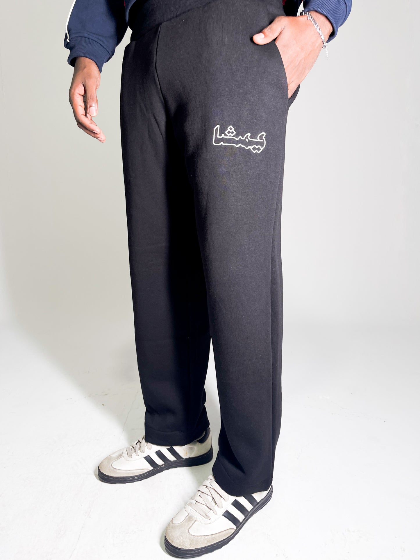 Model wearing black sweatpants with an Arabic print and white sneakers against a light gray background