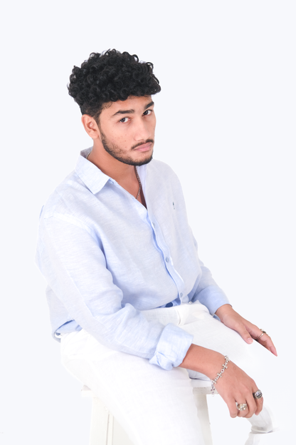 Male model seated on a stool wearing Version Originale’s classic blue linen shirt with navy palm tree embroidery, paired with Version Originale’s white linen pants