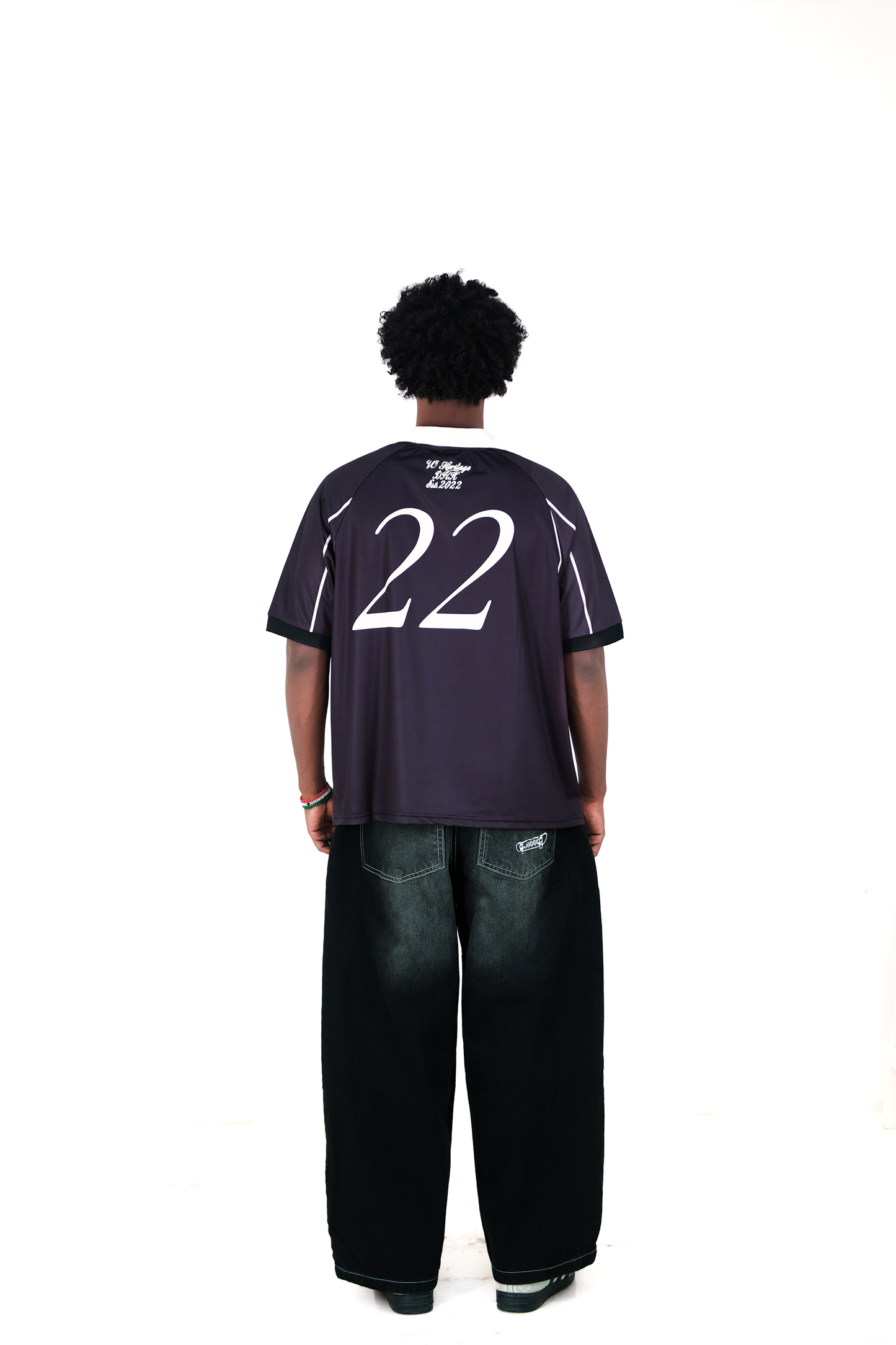Back view of a male model wearing Version Originale’s black athletic jersey with beige collar and piping, featuring the number 22 and 'VO Heritage DHK Est. 2022' printed on the upper back, paired with Version Originale’s wide-leg black faded jeans