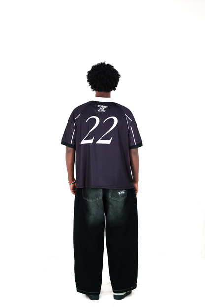 Back view of a male model wearing Version Originale’s black athletic jersey with beige collar and piping, featuring the number 22 and 'VO Heritage DHK Est. 2022' printed on the upper back, paired with Version Originale’s wide-leg black faded jeans